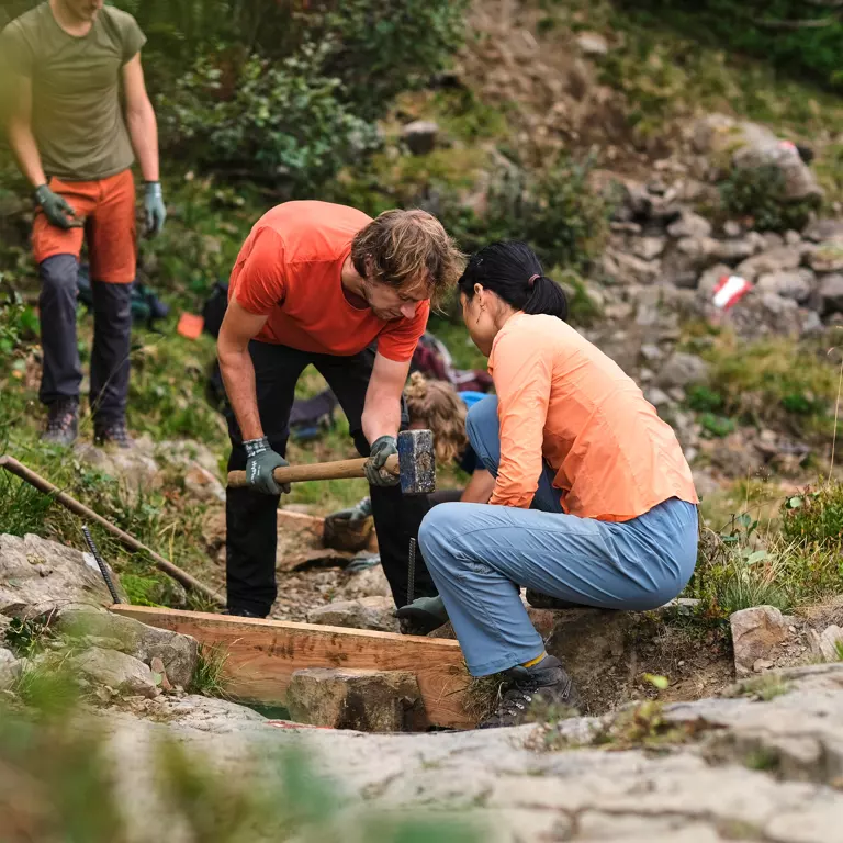 Twee mensen met handschoenen aan en in vrijetijdskleding werken samen op een rotsachtig pad, waarbij ze met gereedschap een houten constructie bouwen of repareren. Op de achtergrond zijn andere mensen en groen te zien. Twee mensen met handschoenen aan en in vrijetijdskleding werken samen op een rotsachtig pad, waarbij ze met gereedschap een houten constructie bouwen of repareren. Op de achtergrond zijn andere mensen en groen te zien.
